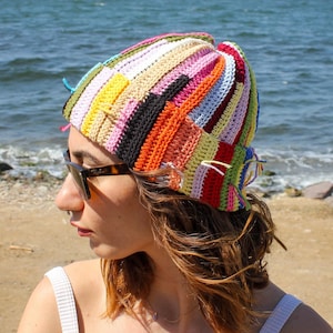 May include: A woman wearing a colourful crocheted beanie hat with a striped pattern. The hat is made with a variety of colours, including pink, orange, yellow, green, blue, and purple. The woman is wearing sunglasses and has long brown hair.