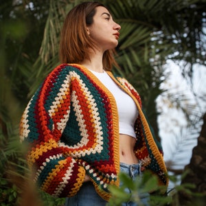 May include: A woman wearing a colorful crocheted cardigan with a white tank top and blue jeans. The cardigan has a striped pattern with red, orange, yellow, green, and white.