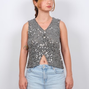 Può includere: Un gilet senza maniche argentato con paillettes, scollo a V e chiusura con bottoni. Il gilet è adornato da numerose paillettes argentate, creando un effetto scintillante. È abbinato a jeans di denim blu chiaro.
