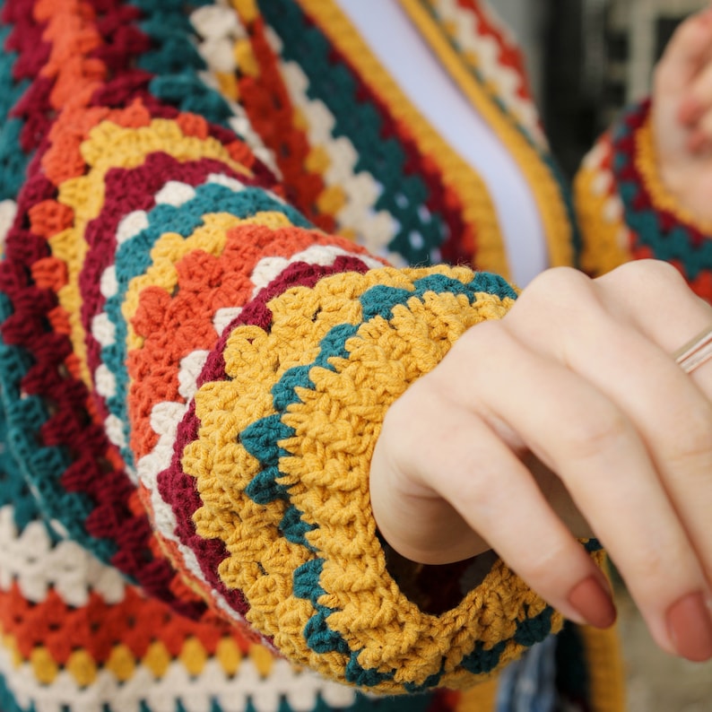 May include: A close-up of a colorful crocheted cardigan with a yellow, teal, orange, red, and white pattern. The cardigan is made with a chunky yarn and has a loose, relaxed fit.