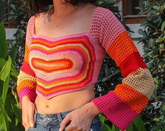 May include: A vibrant, hand-crocheted crop top with a heart motif in pink, orange, and beige. It has long sleeves with flared cuffs and a square neckline. The top is a striking piece of handmade clothing.
