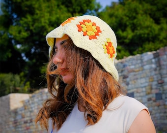 Può includere: Un cappello a secchiello all'uncinetto color crema con disegni floreali arancioni e gialli. Il cappello ha una tesa e viene indossato all'aperto. Lo sfondo comprende alberi verdi e un muro di pietra.