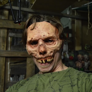 May include: A realistic, flesh-toned zombie mask with exposed teeth and a decaying appearance. The mask covers the face and is worn by a person wearing a green shirt. The background is a dimly lit, rustic interior.