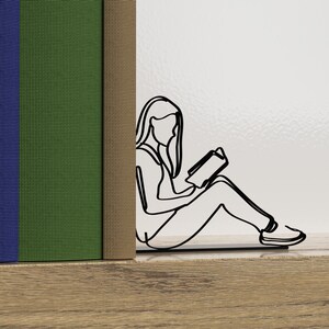 Op de afbeelding: Een zwarte metalen boekenlegger in de vorm van een persoon die zit en een boek leest. De boekenlegger houdt twee boeken vast, een blauwe, een groene en een bruine.