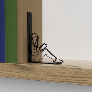 Op de afbeelding: Zwarte metalen boekenleggers in de vorm van een persoon die zit en een boek leest. De boekenleggers staan op een houten plank met een stapel boeken erachter.