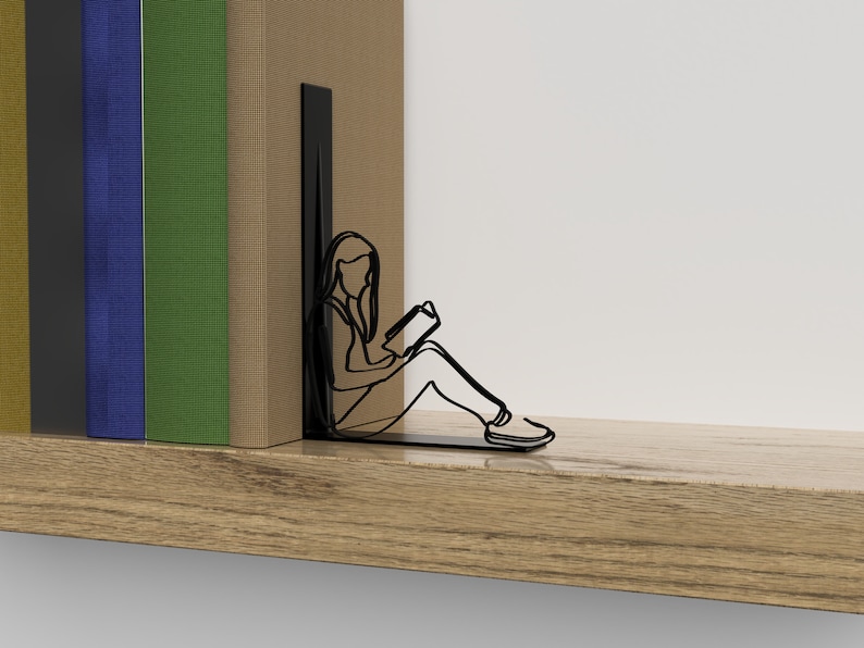 Op de afbeelding: Zwarte metalen boekenleggers in de vorm van een persoon die zit en een boek leest. De boekenleggers staan op een houten plank met een stapel boeken erachter.