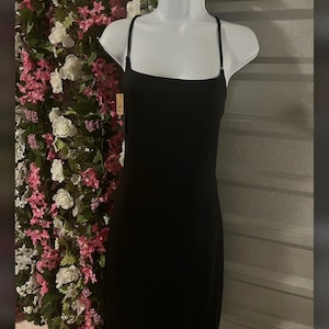 Puede incluir: Un minivestido negro con tirantes finos se muestra en un maniquí blanco. El vestido tiene un escote cuadrado y una silueta ajustada. El fondo presenta un arreglo floral con flores rosas y blancas.