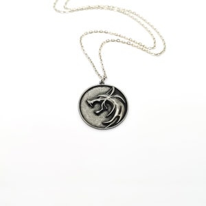 Può includere: Una collana con catena d'argento con un ciondolo rotondo che presenta un design a testa di lupo.
