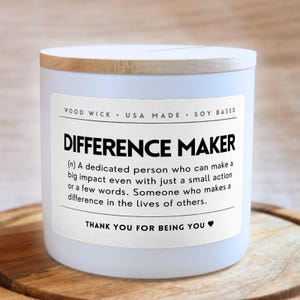May include: A white soy-based candle with a wooden lid. The label reads "DIFFERENCE MAKER" with a definition and "THANK YOU FOR BEING YOU." The candle is USA made and has a wood wick.