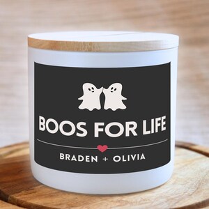 Può includere: Candela bianca con un'etichetta nera che dice "BOOS FOR LIFE" con due illustrazioni di fantasmi e i nomi "BRADEN + OLIVIA" sotto.