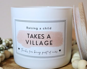 May include: White candle with a black and white label that reads "Raising a child takes a village. Thanks for being part of ours." The label has a pink watercolor background.