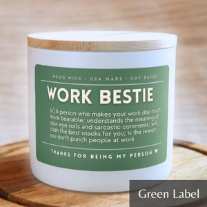 May include: White candle with a green label that reads "Work Bestie" and a definition of the term. The candle has a wooden lid and is sitting on a wooden surface.