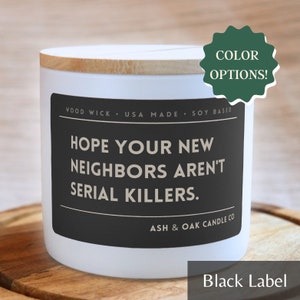Op de afbeelding: Witte kaars met een zwart label waarop staat: "Hopelijk zijn je nieuwe buren geen seriemoordenaars." Het label vermeldt ook "Wood Wick, USA Made, Soy Based" en "Ash & Oak Candle Co."