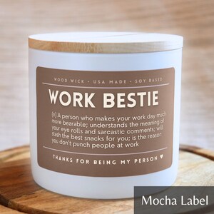 May include: A white candle with a brown label that reads "Work Bestie" and a definition of the term. The label also says "Wood Wick, USA Made, Soy Based" and "Thanks for being my person". The candle has a wooden lid.