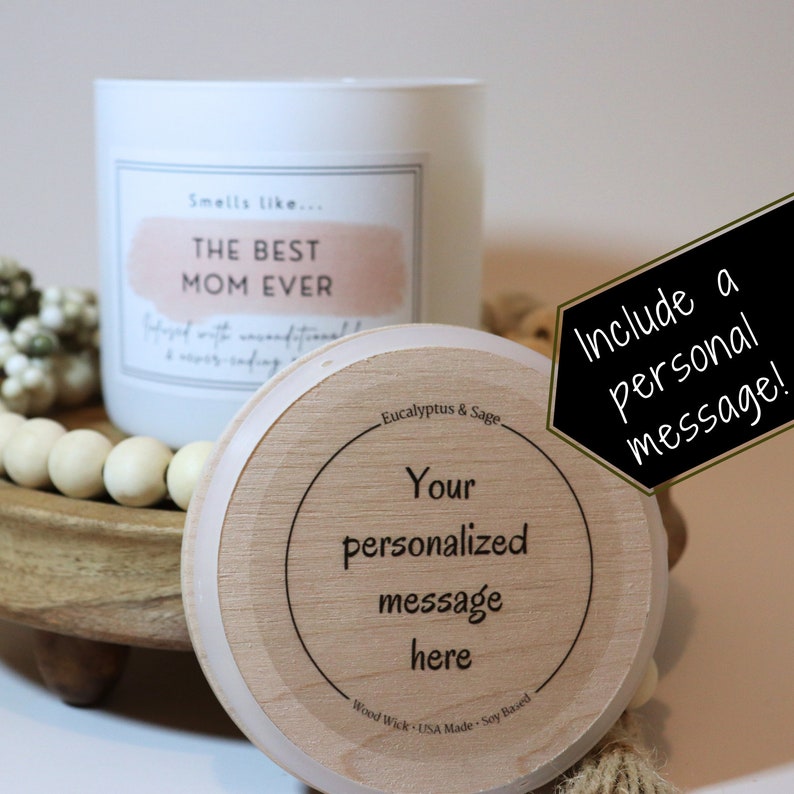 May include: A white candle with a wooden lid. The candle label reads "Smells like... THE BEST MOM EVER". The lid is made of wood and has the text "Eucalyptus & Sage" and "Your personalized message here" printed on it. The candle is on a wooden tray with white beads.