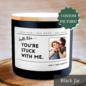 Op de afbeelding: Een zwarte kaars pot met een houten deksel. Het etiket luidt "Smells like... You're stuck with me." en bevat een foto van een stel. De tekst "Wood Wick - USA Made - Soy Based" en "Ash & Oak Candle Co" staat ook op het etiket.
