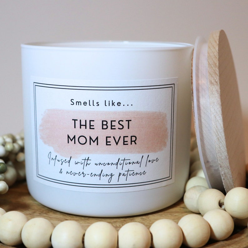 May include: White candle with a pink and white label that reads "Smells like... THE BEST MOM EVER Infused with unconditional love & never-ending patience". The candle has a wooden lid.