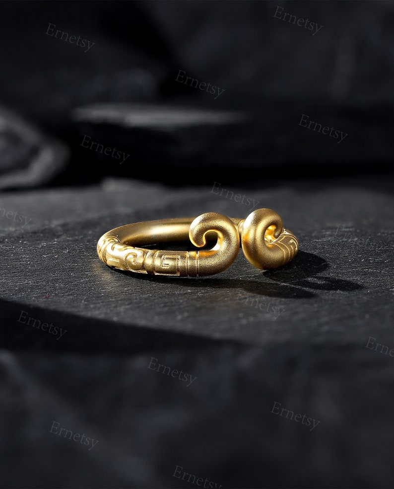 May include: A gold-colored ring with a unique design. The ring features a curved shape with decorative swirls and patterns. The ring is displayed on a dark, textured surface, highlighting its intricate details and craftsmanship.