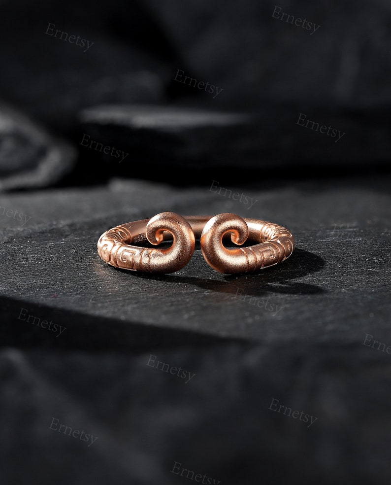 May include: A rose gold ring with a unique design, featuring two swirling ends. The ring is set against a dark, textured surface, highlighting its intricate details and warm color.