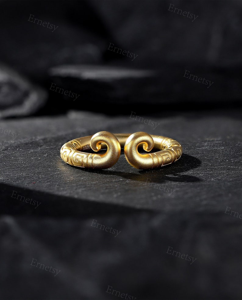 May include: A gold-colored ring with a unique design. The ring features two swirling, scroll-like ends connected by a band with etched details. The ring is set against a dark, textured surface, highlighting its metallic sheen.