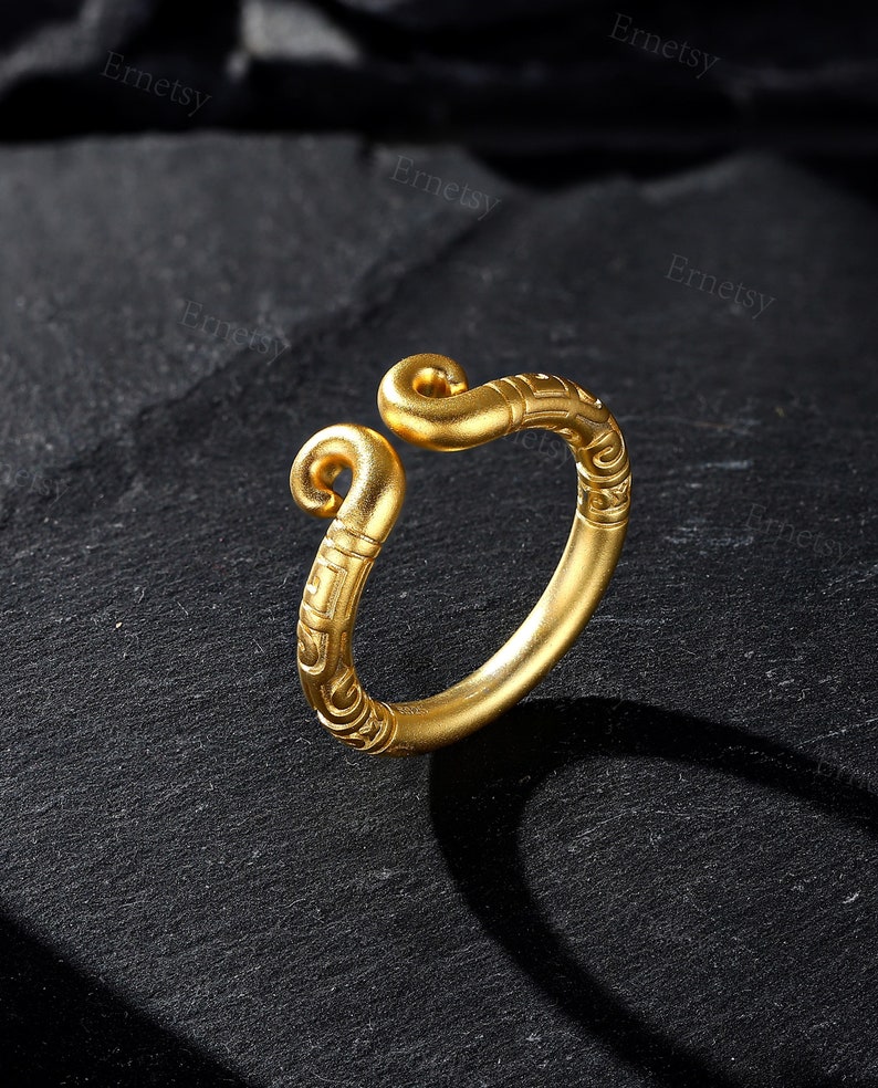 May include: A gold-colored ring with an open design. The ring features intricate engraved patterns and two curled ends. The ring is displayed on a dark, textured surface.