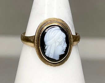Op de afbeelding: Goudkleurige ring met een ovale camee. De camee toont een wit profiel tegen een zwarte achtergrond. De ringband en zetting hebben een vintage uitstraling. De camee toont een klassieke figuur.