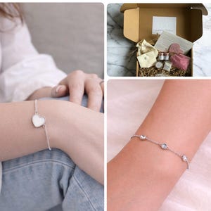 May include: Silver bracelets are shown, one with a white heart charm, the other with three clear gemstones. A gift box contains small bottles, a pink cloth, and a small bag. The bracelets are delicate.