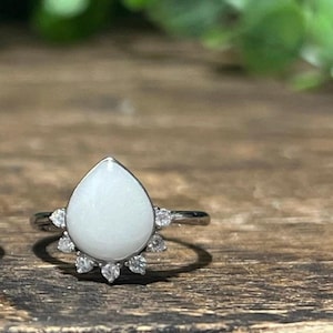 K&ouml;nnte beinhalten: Ein silberner Ring mit einem birnenf&ouml;rmigen, undurchsichtigen wei&szlig;en Stein. Der Stein ist von einem Kranz aus kleinen, klaren Edelsteinen umgeben. Der Ring liegt auf einer Holzoberfl&auml;che mit verschwommenem gr&uuml;nem Laub im Hintergrund.