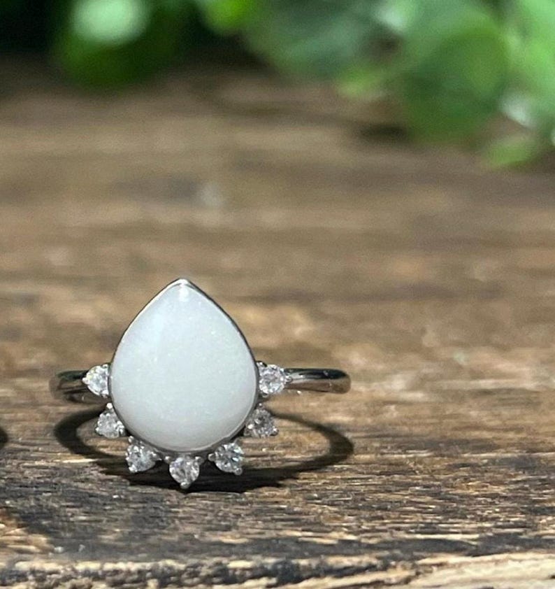 K&ouml;nnte beinhalten: Ein silberner Ring mit einem birnenf&ouml;rmigen, undurchsichtigen wei&szlig;en Stein. Der Stein ist von einem Kranz aus kleinen, klaren Edelsteinen umgeben. Der Ring liegt auf einer Holzoberfl&auml;che mit verschwommenem gr&uuml;nem Laub im Hintergrund.
