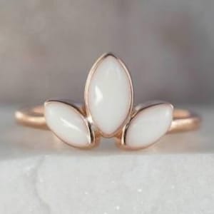 K&ouml;nnte beinhalten: Ein ros&eacute;goldener Ring mit drei wei&szlig;en, marquisef&ouml;rmigen Steinen. Die Steine sind in einem kronenartigen Design angeordnet, wobei der zentrale Stein etwas gr&ouml;&szlig;er ist. Der Ring wird auf einer wei&szlig;en Oberfl&auml;che pr&auml;sentiert.