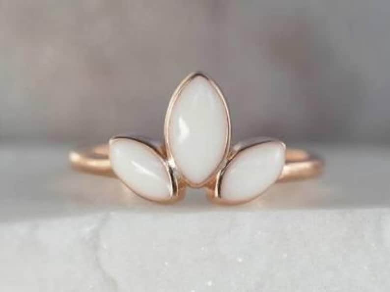 K&ouml;nnte beinhalten: Ein ros&eacute;goldener Ring mit drei wei&szlig;en, marquisef&ouml;rmigen Steinen. Die Steine sind in einem kronenartigen Design angeordnet, wobei der zentrale Stein etwas gr&ouml;&szlig;er ist. Der Ring wird auf einer wei&szlig;en Oberfl&auml;che pr&auml;sentiert.