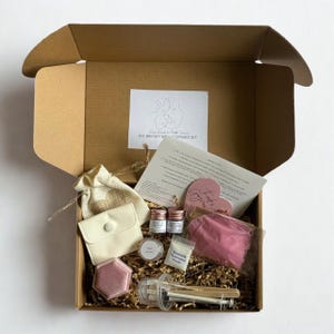 K&ouml;nnte beinhalten: Offener Karton mit einem DIY-Muttermilch-Andenken-Kit. Das Kit enth&auml;lt kleine Gl&auml;ser, einen Beutel, eine herzf&ouml;rmige rosa Karte, ein kleines sechseckiges rosa Objekt und eine Spritze. Die Box enth&auml;lt auch einen kleinen Beutel und eine wei&szlig;e Karte mit Anweisungen.