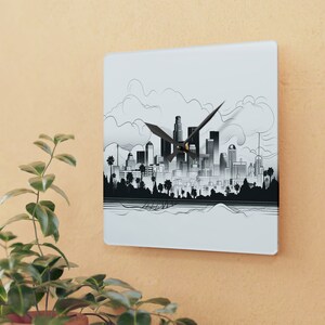 May include: Square wall clock with a black and white city skyline illustration. The clock hands are black. The design includes buildings, trees, and clouds. The clock is mounted on a light orange wall.