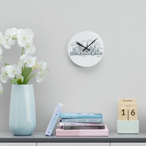 May include: A white round clock with a black outline of a city skyline. The clock hands are black and the clock face is white. The clock is hanging on a white wall. A wooden calendar block with the date July 16th is on a shelf with a stack of books.