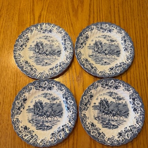 Op de afbeelding: Vier bijpassende blauw-witte decoratieve borden. Elk bord heeft een gedetailleerde illustratie van een paardenkoetsscène. De borden hebben een geschulpte rand met een bloemmotief. De borden zijn gerangschikt op een houten oppervlak.