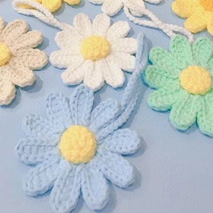 May include: A collection of crocheted daisy flowers in various colors, including white, light blue, light green, and cream. The flowers are attached to strings, suggesting they are ornaments or charms.