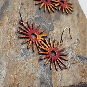 May include: A pair of red and yellow sunburst earrings with silver hooks. The earrings have a circular center with radiating lines, creating a star-like design. Additional earrings are visible in the background, all resting on a textured stone surface.