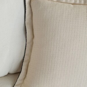 Puede incluir: Primer plano de un coj&iacute;n color crema con una superficie texturizada y un ribete negro. El coj&iacute;n tiene un patr&oacute;n cuadrado y forma parte de una colecci&oacute;n de decoraci&oacute;n del hogar.