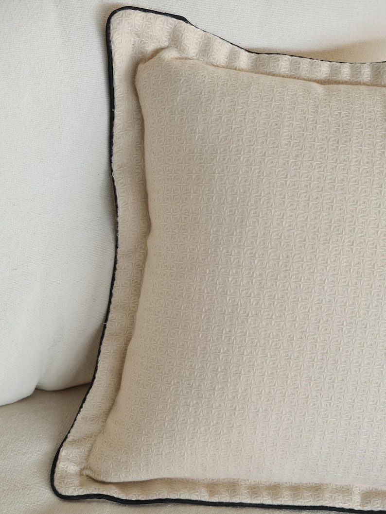 Puede incluir: Primer plano de un coj&iacute;n color crema con una superficie texturizada y un ribete negro. El coj&iacute;n tiene un patr&oacute;n cuadrado y forma parte de una colecci&oacute;n de decoraci&oacute;n del hogar.