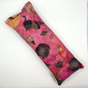 Puede incluir: Una almohada rectangular rosa con temática de Halloween. La almohada presenta ilustraciones de un gato negro con sombrero de bruja, botellas de pociones, un caldero, caramelos y una araña. La almohada tiene un efecto acuarela.