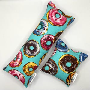 May include: Two cat toys, one long and one square, are displayed. They are covered in a teal fabric printed with colorful donuts. The donuts have pink, brown, yellow, and blue frosting. The label "The Three Twinkies" is visible.