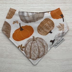 以下が含まれることがあります： オレンジと茶色のカボチャ、葉、そして「The Three Twindlers」という文字が書かれた、秋のテーマの白い布製のバンダナです。