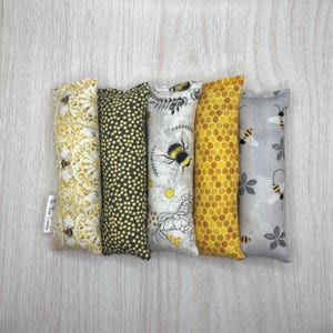Puede incluir: Cinco juguetes de tela para gatos con hierba gatera en forma de almohada larga. Los juguetes están cubiertos con diferentes patrones de temática de abejas, incluyendo panal, abejas y flores. Los juguetes están todos alineados en una fila sobre una superficie de madera clara. El texto "The Meow Trends" está impreso en uno de los juguetes.