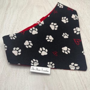 May include: Black and red pet bandana with white paw prints and red hearts. The bandana is reversible, with a solid red interior. A white tag with the text "The Three Tuxedos" is sewn on the bottom.