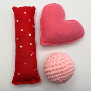 Puede incluir: Tres juguetes de peluche con temática de San Valentín: una almohada roja con corazones blancos, una almohada rosa en forma de corazón y una bola rosa de ganchillo.