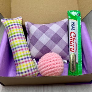 Puede incluir: Una caja de cartón llena de juguetes para gatos. Los juguetes incluyen una almohada a cuadros morado y blanco, una pelota de crochet rosa, un juguete a cuadros multicolor y un paquete de comida para gatos verde y blanco etiquetado como "INABA Churu - ¡Irresistible! Atún con receta de pollo".