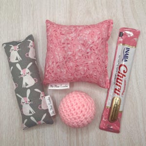 Puede incluir: Un conjunto de juguetes para gatos, que incluye una almohada de hierba gatera gris con estampado de conejos, una almohada rosa floral, una bola rosa de ganchillo y una golosina Churu rosa y blanca. Etiqueta The Three Tuxedos.