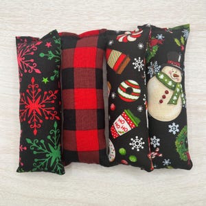Puede incluir: Cuatro juguetes de hierba gatera festivos con diferentes patrones navideños. Los juguetes son negros con acentos rojos y verdes. Un juguete tiene un muñeco de nieve, otro tiene bastones de caramelo, otro tiene copos de nieve y el último tiene un patrón de cuadros de búfalo.
