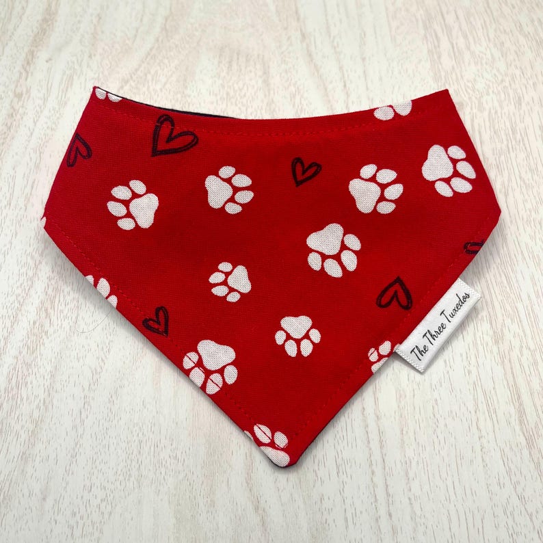 May include: Red dog bandana with white paw prints and black hearts. The bandana is a triangle shape and has a tag that says "The Three Tuxedos". The bandana is made of fabric.