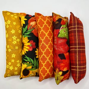 Puede incluir: Seis juguetes para gatos con diferentes diseños de tela. Los diseños incluyen flores amarillas, girasoles, un diseño geométrico naranja, flores rojas y naranjas y un estampado de cuadros rojos. Cada juguete tiene forma rectangular y alargada.